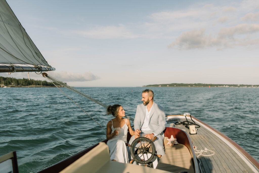 Couple laughing together while groom drives the sailboat and bride drinks wine